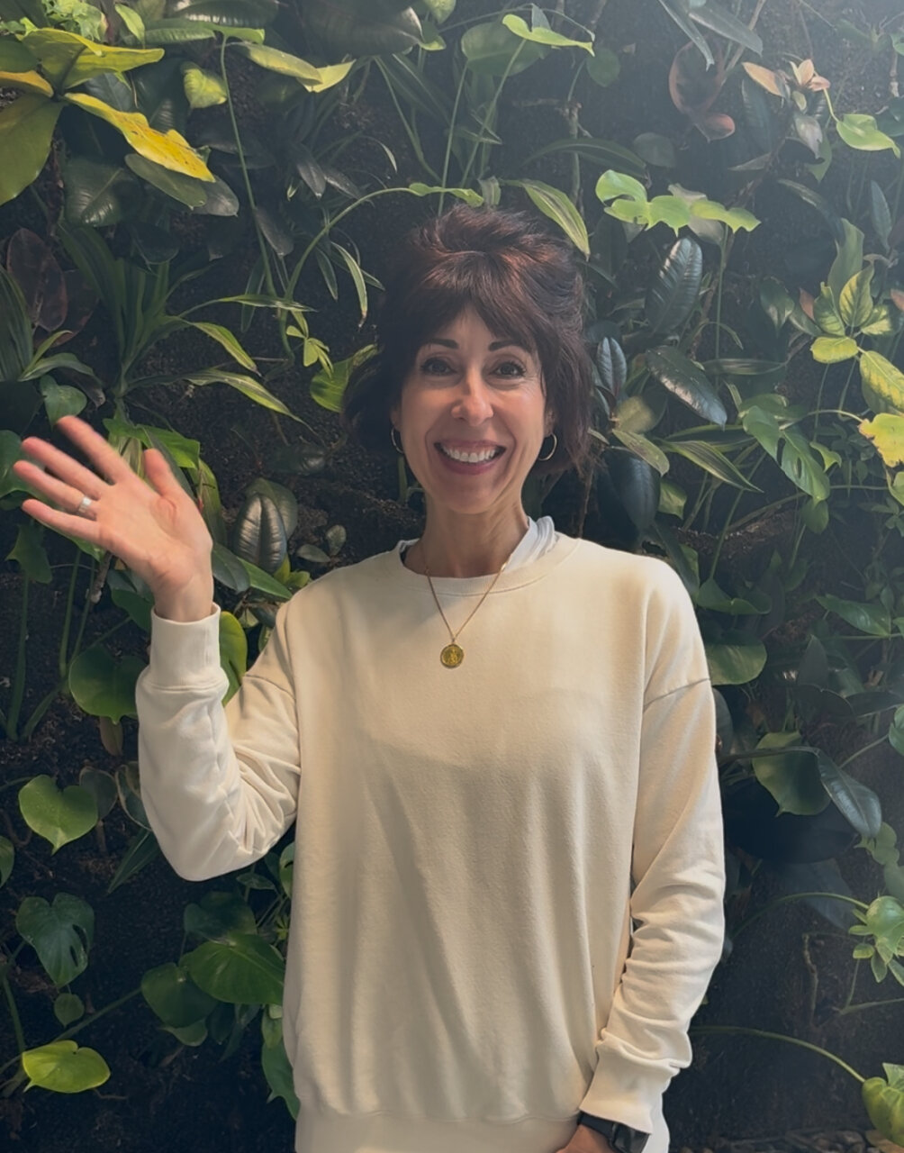 middle aged woman standing and waving in front of a live wall of green leafy plants